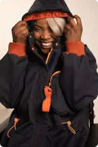 a smiling woman sitting down wears a black hooded coat with orange trim